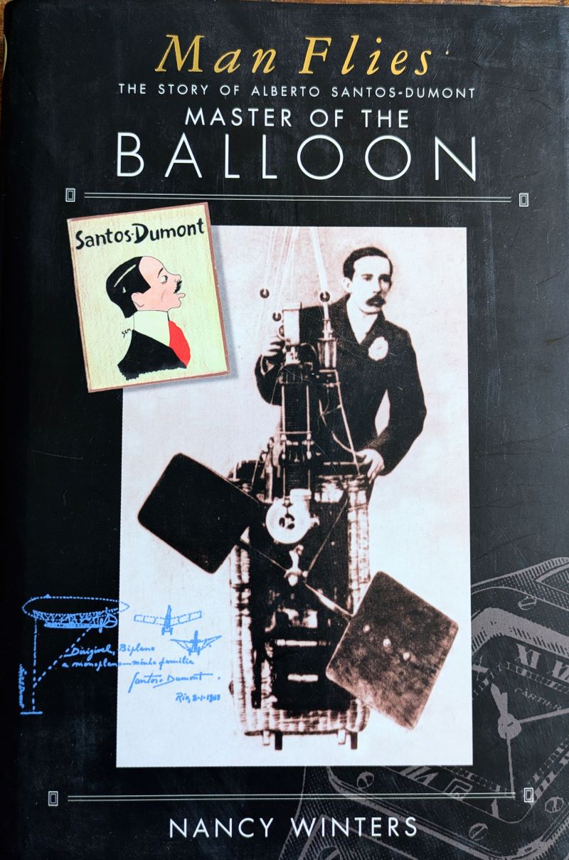 Picture of colour pictorial book cover showing Santos-Dumont standing in a small basket with primitive motor and propeller attached to the front.